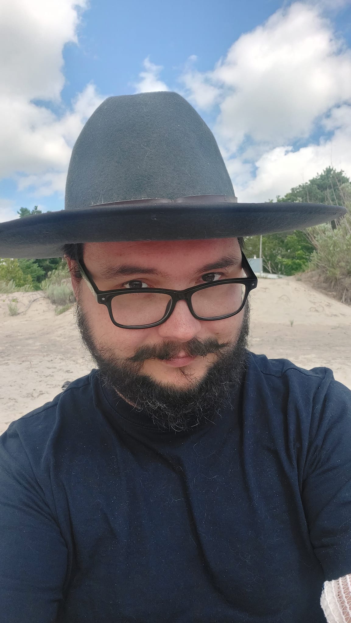 A man short hair and bushy facial hair wears a wide brimmed felt sunhat on a beach with a sky filled with fluffy white clouds